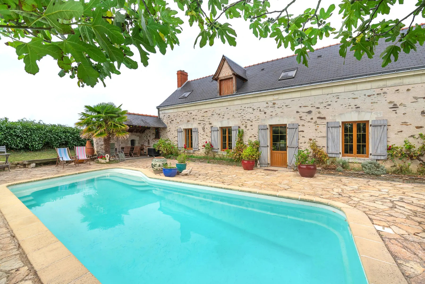 Maison en pierre aux volets bleus derrière une piscine bordée de dalles